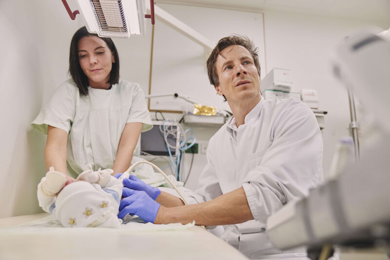 Das Bild zeigt einen Arzt und eine Mitarbeiterin bei der Untersuchung eines Neugeborenen.